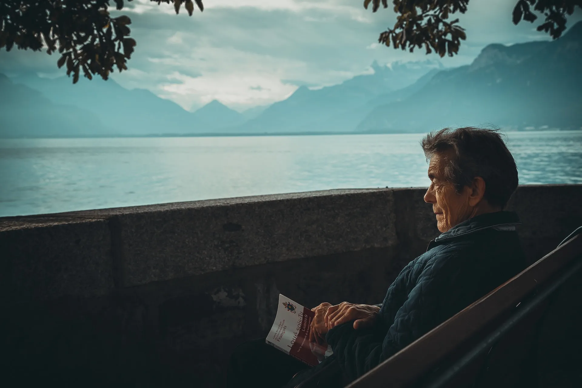 An elderly man, with gray hair and glasses, engrossed in reading a book while seated on a wooden bench.