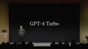 A man on stage, with a projector screen behind him. ChatGPT-4 Turbo Event.